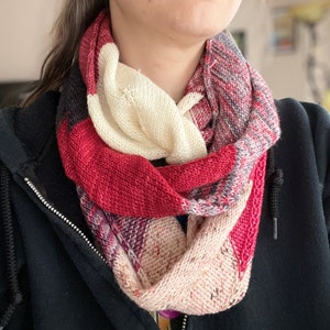 Puede incluir: Una bufanda de punto multicolor con tonos de rojo, blanco, rosa y negro. La bufanda está doblada y se lleva alrededor del cuello.