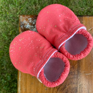 Op de afbeelding: Een paar roze babyschoentjes met gouden stippen. De schoentjes hebben een witte rand en zijn gemaakt van zacht materiaal.