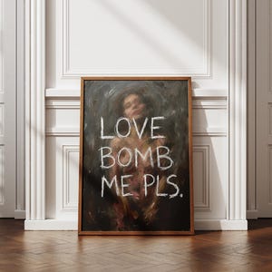 Könnte beinhalten: Ein gerahmtes Kunstwerk mit dunklem Hintergrund und einem verschwommenen Bild einer Person. Die Worte "LOVE BOMB ME PLS." sind in weißen, kreideartigen Buchstaben geschrieben. Der Rahmen hat eine warme braune Farbe, und das Kunstwerk wird vor einer weißen Wand ausgestellt.