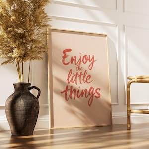 Könnte beinhalten: Ein gerahmter Druck mit den Worten "Enjoy the little things" in roter Schrift. Der Druck lehnt an einer weißen Wand neben einer großen, dunkelbraunen Vase mit getrocknetem Pampasgras. Im Hintergrund steht ein kleiner Beistelltisch aus Bambus.