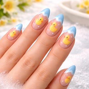 May include: A hand with long, almond-shaped nails. Each nail features a light pink base with a blue tip and a yellow duck design. White dots accent the duck design, creating a playful and whimsical aesthetic.
