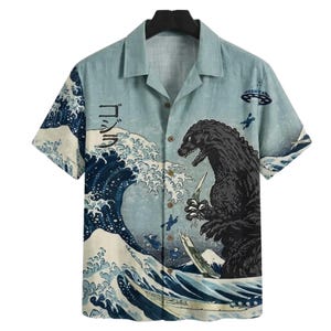 May include: Light blue button-up shirt featuring a Godzilla design. The shirt showcases a Japanese wave pattern with Godzilla and a UFO. The Japanese word "ゴジラ" is printed on the shirt.