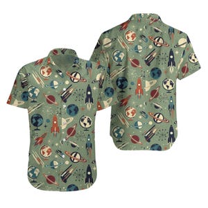 May include: Short-sleeved button-up shirt with a retro space-themed pattern. The design features rockets, planets, globes, and starbursts in shades of red, blue, and white on a green background. The shirt has a classic collar and button closure.