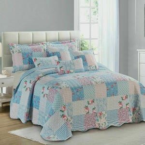May include: A bedspread set with a patchwork design in blue, pink, and white, with floral and geometric patterns. The set includes a quilt and matching pillow shams. The bed has a white headboard and a matching bedside table.