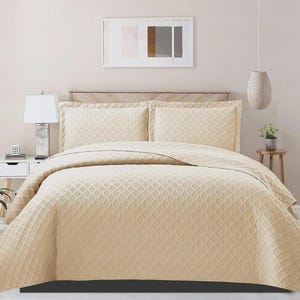 May include: A beige bedspread and two matching pillowcases with a geometric pattern. The bed has a wooden headboard. A white lamp sits on a bedside table to the left. A decorative hanging light and a small plant are also visible.