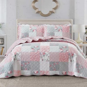 May include: A patchwork quilt and two pillow shams in pink, grey, and white. The quilt features floral prints, gingham, and solid squares. The bedding set is arranged on a bed with a neutral headboard. The quilt is designed for a double bed.
