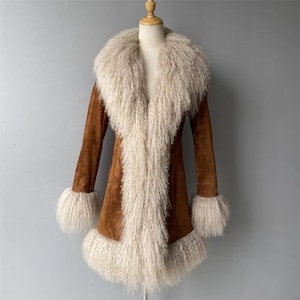 May include: A brown suede coat with a cream-colored, shaggy fur collar, cuffs, and front panel. The coat is displayed on a mannequin. The coat has long sleeves and a mid-thigh length.