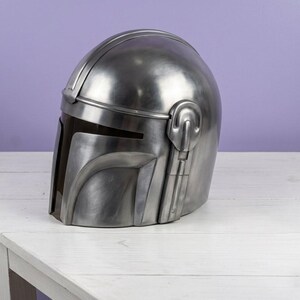K&ouml;nnte beinhalten: Ein metallisch silberner Helm mit einer glatten, abgerundeten Kuppel und einer dunklen, horizontalen Visier&ouml;ffnung. Der Helm verf&uuml;gt &uuml;ber ein markantes T-f&ouml;rmiges Frontplattendesign mit erhabenen strukturellen Details an den Seiten und einer polierten Oberfl&auml;che.
