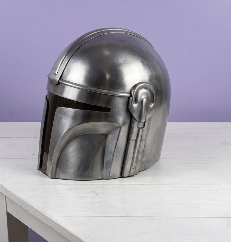 K&ouml;nnte beinhalten: Ein metallisch silberner Helm mit einer glatten, abgerundeten Kuppel und einer dunklen, horizontalen Visier&ouml;ffnung. Der Helm verf&uuml;gt &uuml;ber ein markantes T-f&ouml;rmiges Frontplattendesign mit erhabenen strukturellen Details an den Seiten und einer polierten Oberfl&auml;che.