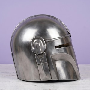 K&ouml;nnte beinhalten: Ein metallisch silberner Helm mit einer glatten, abgerundeten Kuppel und einer T-f&ouml;rmigen Visier&ouml;ffnung. Der Helm verf&uuml;gt &uuml;ber einen markanten, erhabenen Grat entlang der Mitte und ein mechanisches Ohrst&uuml;ck-Detail an der Seite mit gl&auml;nzendem Finish.
