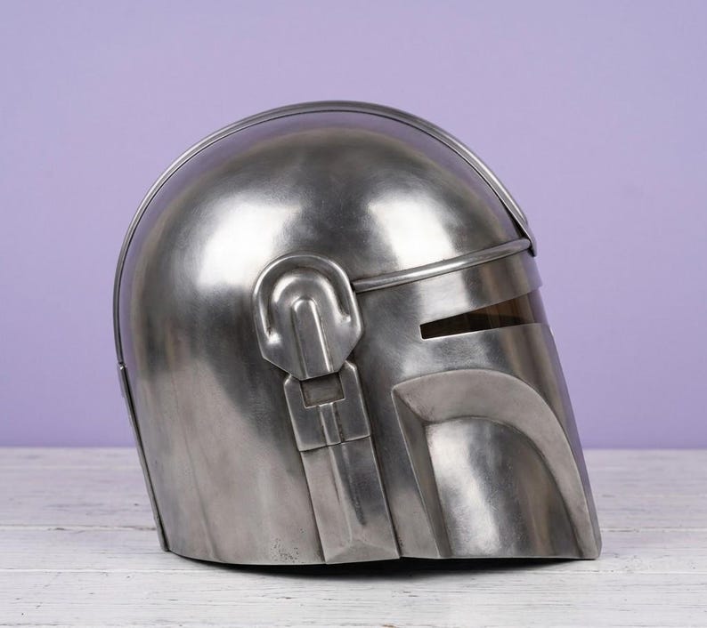K&ouml;nnte beinhalten: Ein metallisch silberner Helm mit einer glatten, abgerundeten Kuppel und einer T-f&ouml;rmigen Visier&ouml;ffnung. Der Helm verf&uuml;gt &uuml;ber einen markanten, erhabenen Grat entlang der Mitte und ein mechanisches Ohrst&uuml;ck-Detail an der Seite mit gl&auml;nzendem Finish.