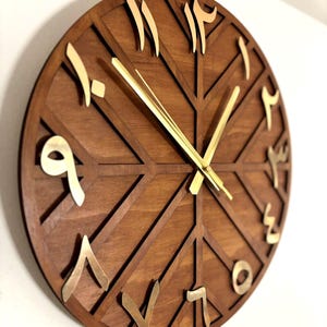 K&ouml;nnte beinhalten: Eine runde Wanduhr aus Holz mit geometrischem Design. Das Zifferblatt ist braun mit goldfarbenen arabischen Ziffern und Zeigern. Das Design der Uhr weist eine Reihe sich kreuzender Linien auf, die ein einzigartiges Muster bilden.