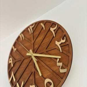 K&ouml;nnte beinhalten: Eine runde Wanduhr aus Holz mit arabischen Ziffern in Gold. Das Zifferblatt hat eine dunkelbraune Farbe mit einem geometrischen Design. Die Zeiger der Uhr sind goldfarben.
