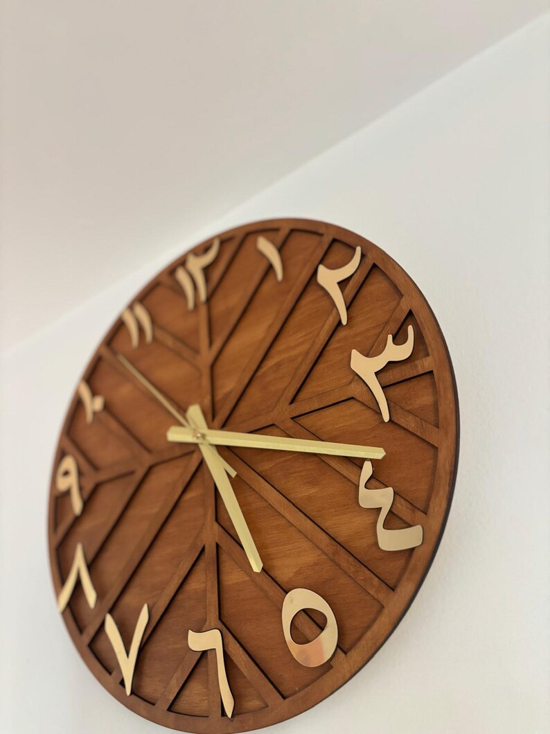 K&ouml;nnte beinhalten: Eine runde Wanduhr aus Holz mit arabischen Ziffern in Gold. Das Zifferblatt hat eine dunkelbraune Farbe mit einem geometrischen Design. Die Zeiger der Uhr sind goldfarben.