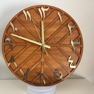 K&ouml;nnte beinhalten: Eine runde Wanduhr aus Holz mit arabischen Ziffern. Das Zifferblatt hat eine warme braune Farbe mit einem geometrischen Design. Die Ziffern und Zeiger sind goldfarben. Die Uhr wird auf einer wei&szlig;en Oberfl&auml;che angezeigt.