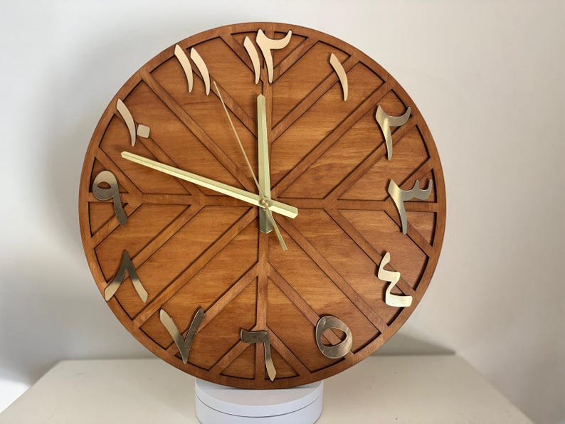 K&ouml;nnte beinhalten: Eine runde Wanduhr aus Holz mit arabischen Ziffern. Das Zifferblatt hat eine warme braune Farbe mit einem geometrischen Design. Die Ziffern und Zeiger sind goldfarben. Die Uhr wird auf einer wei&szlig;en Oberfl&auml;che angezeigt.
