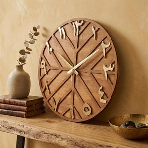 K&ouml;nnte beinhalten: Eine runde Wanduhr aus Holz mit arabischen Ziffern in einem geometrischen Design. Die Uhr hat goldfarbene Zeiger und Ziffern. Sie wird auf einem Holztisch mit einer Vase, B&uuml;chern und einer dekorativen Schale ausgestellt.