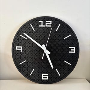 May include: A circular black wall clock featuring white numerals and hands. The clock face has a textured design. White lines mark the hours. The clock is on a white surface.
