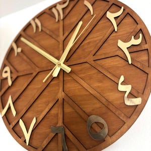 K&ouml;nnte beinhalten: Eine runde Holzuhr mit geometrischem Design. Das Zifferblatt ist in einem warmen Braunton gehalten, mit goldfarbenen arabischen Ziffern und Zeigern. Das Design weist sich kreuzende Linien auf, die ein einzigartiges Muster bilden.