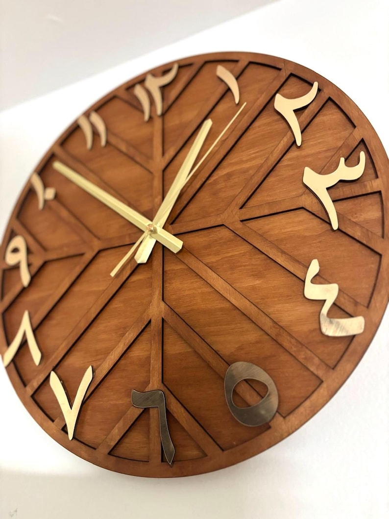 K&ouml;nnte beinhalten: Eine runde Holzuhr mit geometrischem Design. Das Zifferblatt ist in einem warmen Braunton gehalten, mit goldfarbenen arabischen Ziffern und Zeigern. Das Design weist sich kreuzende Linien auf, die ein einzigartiges Muster bilden.