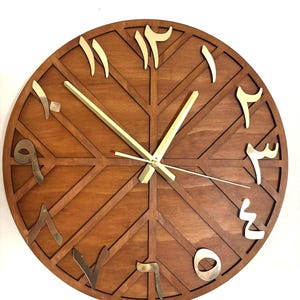 K&ouml;nnte beinhalten: Eine runde Holzuhr mit arabischen Ziffern. Das Zifferblatt der Uhr ist warmbraun mit einem geometrischen Muster. Die Ziffern sind goldfarben und die Zeiger der Uhr sind goldfarben. Die Uhr ist an einer wei&szlig;en Wand befestigt.
