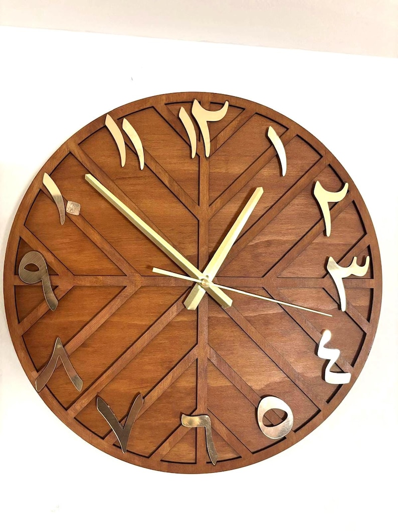 K&ouml;nnte beinhalten: Eine runde Holzuhr mit arabischen Ziffern. Das Zifferblatt der Uhr ist warmbraun mit einem geometrischen Muster. Die Ziffern sind goldfarben und die Zeiger der Uhr sind goldfarben. Die Uhr ist an einer wei&szlig;en Wand befestigt.