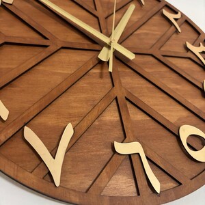 K&ouml;nnte beinhalten: Eine Wanduhr aus Holz mit geometrischem Design. Das Zifferblatt hat eine warme braune Farbe mit goldfarbenen arabischen Ziffern und Zeigern. Das Design weist eine Reihe sich kreuzender Linien auf, die einen einzigartigen und modernen Look erzeugen.