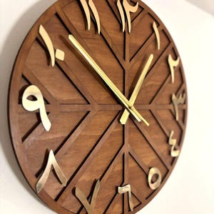 K&ouml;nnte beinhalten: Eine runde Holzuhr mit arabischen Ziffern und goldfarbenen Zeigern. Das Zifferblatt der Uhr ist dunkelbraun mit einem geometrischen Design. Die Ziffern sind goldfarben. Die Uhr ist an einer wei&szlig;en Wand befestigt.