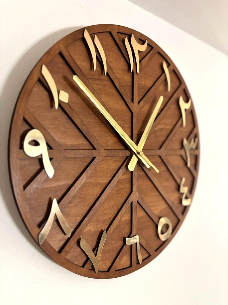 K&ouml;nnte beinhalten: Eine runde Holzuhr mit arabischen Ziffern und goldfarbenen Zeigern. Das Zifferblatt der Uhr ist dunkelbraun mit einem geometrischen Design. Die Ziffern sind goldfarben. Die Uhr ist an einer wei&szlig;en Wand befestigt.