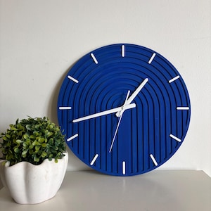 May include: A contemporary blue wall clock featuring white hands and hour indicators. The clock has a distinctive, layered design and is positioned beside a small, green plant in a white pot. The clock's diameter is approximately 30 cm.