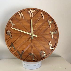 K&ouml;nnte beinhalten: Eine runde Holzuhr mit arabischen Ziffern in Gold. Das Zifferblatt ist hellbraun mit einem geometrischen Design. Die Zeiger der Uhr sind goldfarben. Die Uhr steht auf einer wei&szlig;en Oberfl&auml;che.
