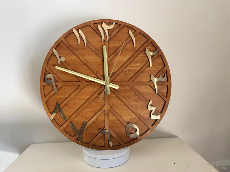 K&ouml;nnte beinhalten: Eine runde Holzuhr mit arabischen Ziffern in Gold. Das Zifferblatt ist hellbraun mit einem geometrischen Design. Die Zeiger der Uhr sind goldfarben. Die Uhr steht auf einer wei&szlig;en Oberfl&auml;che.