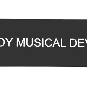May include: A rectangular black sign featuring the white text KOGOY MUSICAL DEVICES in a clean, sans-serif font. The sign has a smooth, matte finish and is oriented horizontally, presenting the brand name clearly against the dark background.