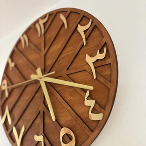 K&ouml;nnte beinhalten: Eine runde Wanduhr aus Holz mit geometrischem Design. Das Zifferblatt ist dunkelbraun mit goldfarbenen arabischen Ziffern und Zeigern. Die Uhr ist an einer wei&szlig;en Wand befestigt.