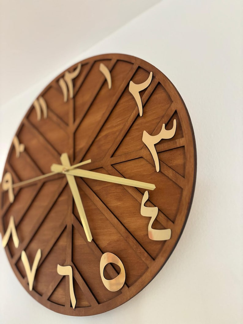 K&ouml;nnte beinhalten: Eine runde Wanduhr aus Holz mit geometrischem Design. Das Zifferblatt ist dunkelbraun mit goldfarbenen arabischen Ziffern und Zeigern. Die Uhr ist an einer wei&szlig;en Wand befestigt.