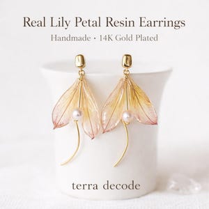 May include: Real Lily Petal Resin Earrings. Handmade 14K Gold Plated earrings featuring delicate, translucent lily petals in shades of pink and yellow. Each earring has a small pearl and gold-coloured accents. The earrings are displayed on a white background.