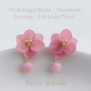 May include: Pink hydrangea resin earrings with 14k gold-plated accents. Each earring features a delicate flower design with a small pink bead. The earrings are handmade and have a floral theme.