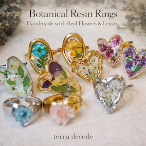 以下が含まれることがあります： 本物の花と葉を使用した、ボタニカルレジンリングのコレクション。リングは金と銀のバンドで、ハート型と楕円型のレジンセッティングが施されています。花は青、黄色、ピンク、紫など、さまざまな色があります。