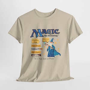 Puede incluir: Camiseta beige con el texto azul "MAGIC The Gathering" y una ilustración de mago. La camiseta también enumera las fases del juego y la frase "It's Not Just a Phase".
