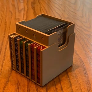 Può includere: Un organizer in legno per carte e dadi con esterno beige chiaro e interno in legno naturale. L'organizer ha scomparti per dadi di vari colori e una sezione più grande per le carte. Gli scomparti dei dadi sono numerati.