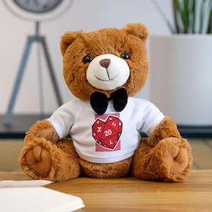 May include: A brown teddy bear wearing a white t-shirt with a red heart-shaped graphic featuring a die with the number 20. The bear has a black bow tie and is sitting on a wooden surface. The bear is a plush toy.