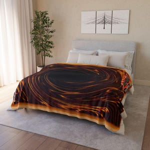Puede incluir: Una manta suave con un diseño arremolinado y ardiente en tonos negros, marrones y dorados. La manta está colocada sobre una cama con ropa de cama blanca. Un logotipo es visible en la esquina.