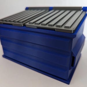 May include: A blue rectangular container with a gray, ridged lid. The container has a boxy shape with horizontal lines and a small opening on the side. The lid is divided into two sections and has a hinged design.