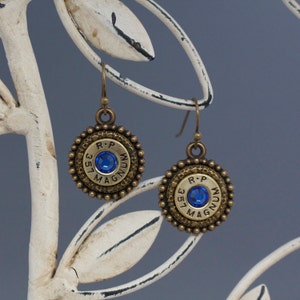 May include: A pair of gold-toned earrings featuring a blue gemstone set in a round bullet casing with the inscription "R-P 357 MAGNUM".