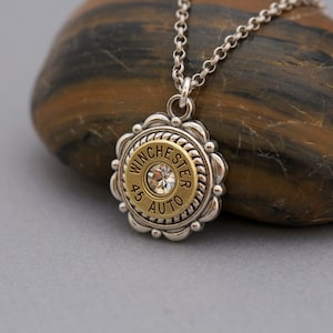 May include: A silver chain necklace with a gold-toned pendant featuring a round bullet casing with the inscription "Winchester 45 Auto" and a clear rhinestone in the center.