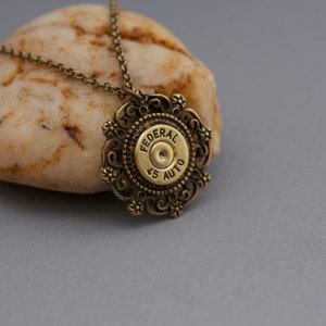 May include: A gold necklace with a pendant featuring a 45 caliber bullet casing with the words "FEDERAL 45 AUTO" inscribed on it. The bullet casing is surrounded by a decorative filigree design.