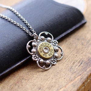 May include: Silver-toned pendant necklace with a floral design. The pendant features a brass-colored center with the text "WINCHESTER 45 AUTO". The necklace is displayed on a wooden surface.
