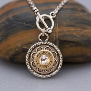 May include: A silver chain necklace with a toggle clasp and a round pendant featuring a gold-colored bullet casing with a clear rhinestone. The bullet casing is engraved with "Hornady 9mm Luger".