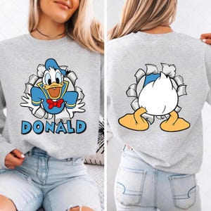 May include: A light gray sweatshirt featuring a cartoon image of Donald Duck bursting through a torn background. The front of the sweatshirt displays Donald Duck in his classic outfit with the name "DONALD" below. The back shows Donald's rear.