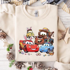 Può includere: Felpa beige con una grafica colorata di auto animate decorate per Pasqua. Le auto includono un'auto da corsa rossa, un'auto blu, un carro attrezzi marrone e un'auto gialla, tutte con decorazioni pasquali e uova di Pasqua.